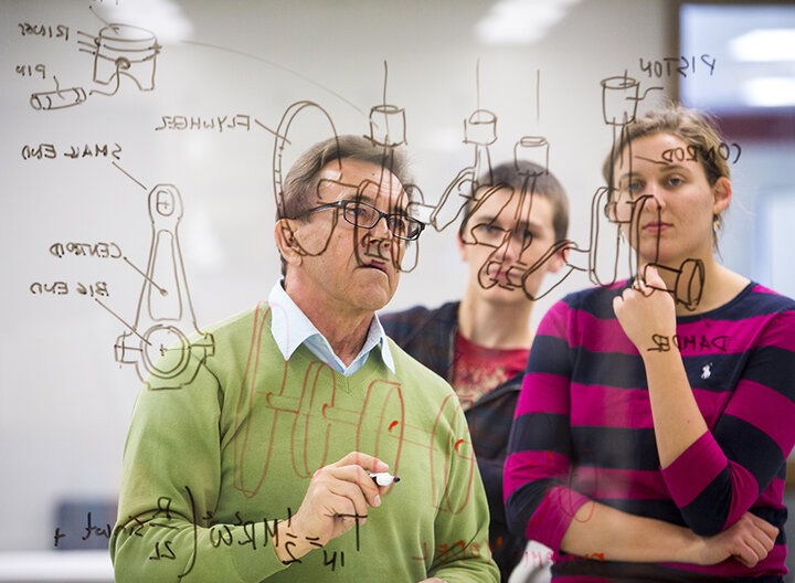 A professor and students stand in front of whiteboard covered in notes.